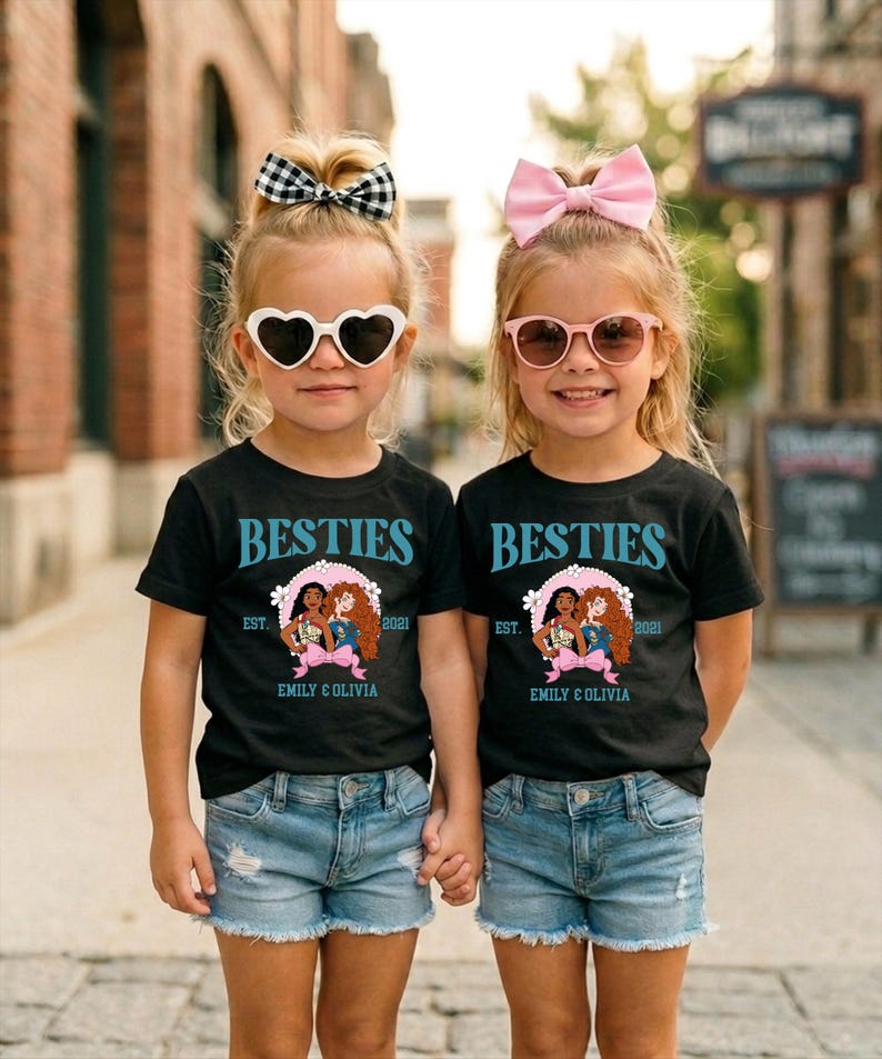 Two young girls wearing matching black t-shirts with the text "BESTIES" and "BESTIES" printed on them, along with a heart-shaped logo, are standing together on a city street.