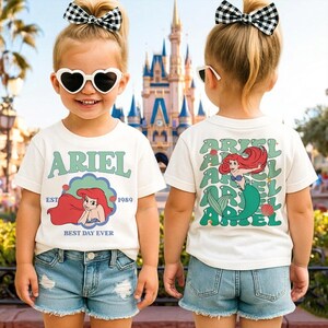 A young girl wearing a white t-shirt with a graphic of Ariel from the Disney movie "The Little Mermaid" and a heart-shaped pair of sunglasses, standing in front of a Disney castle.
