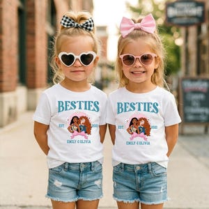 Two young girls wearing matching white t-shirts with the text "BESTIES" and "EST. 2021 EMILY & OLIVIA" printed on them, along with heart-shaped sunglasses and pink bows.