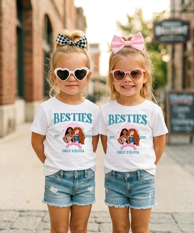Two young girls wearing matching white t-shirts with the text "BESTIES" and "EST. 2021 EMILY & OLIVIA" printed on them, along with heart-shaped sunglasses and pink bows.