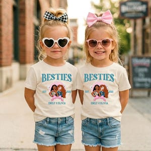 Two young girls wearing matching white t-shirts with the text "BESTIES" and "EST. 2021 EMILY & OLIVIA" printed on them, along with heart-shaped sunglasses and pink bows.