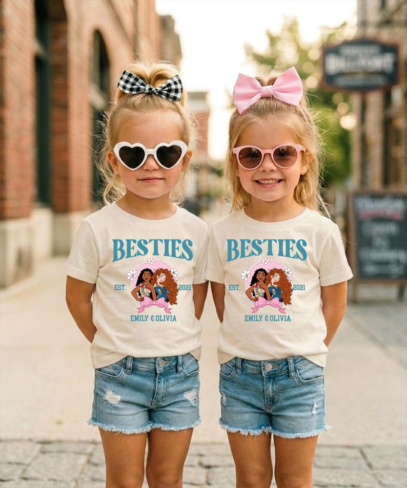 Two young girls wearing matching white t-shirts with the text "BESTIES" and "EST. 2021 EMILY & OLIVIA" printed on them, along with heart-shaped sunglasses and pink bows.