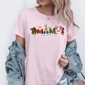 A woman wearing a pink t-shirt with the word "MAMMA" printed on it, along with various colorful cartoon characters. She is also wearing a denim jacket and jeans.