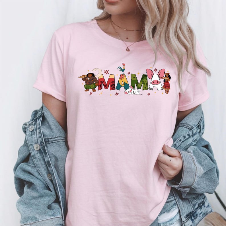 A woman wearing a pink t-shirt with the word "MAMMA" printed on it, along with various colorful cartoon characters. She is also wearing a denim jacket and jeans.