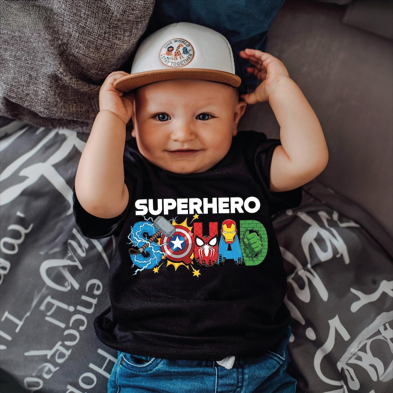 A smiling baby wearing a black t-shirt with superhero designs and a white baseball cap.