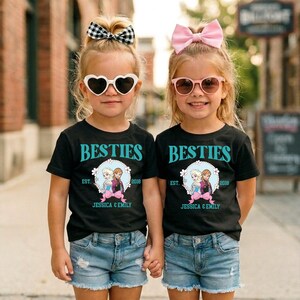 Two young girls wearing matching black t-shirts with the text "BESTIES" and "EST. 2019 JESSICA & EMILY" printed on them, along with heart-shaped sunglasses and pink bows, standing together on a city street.