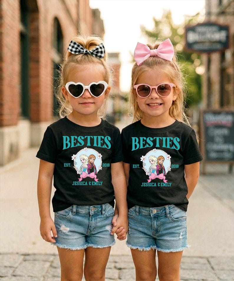 Two young girls wearing matching black t-shirts with the text "BESTIES" and "EST. 2019 JESSICA & EMILY" printed on them, along with heart-shaped sunglasses and pink bows, standing together on a city street.