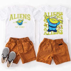 a pair of children's clothing items, including a white t-shirt with a yellow alien character and a pair of brown shorts.