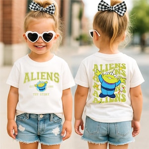 A young girl wearing a white t-shirt with a cartoon character and the text "Aliens" on it, along with a bow in her hair. She is also wearing sunglasses and denim shorts.