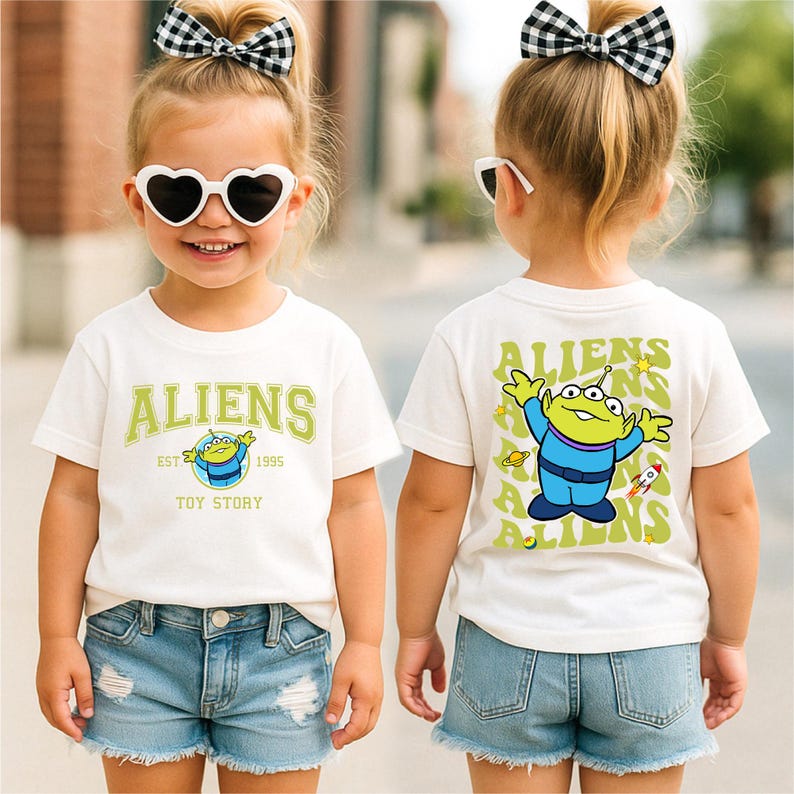 A young girl wearing a white t-shirt with a cartoon character and the text "Aliens" on it, along with a bow in her hair. She is also wearing sunglasses and denim shorts.