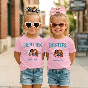 Two young girls wearing matching pink t-shirts with the text "BESTIES" and "EST. 2021 EMILY & OLIVIA" standing side by side on a city street.