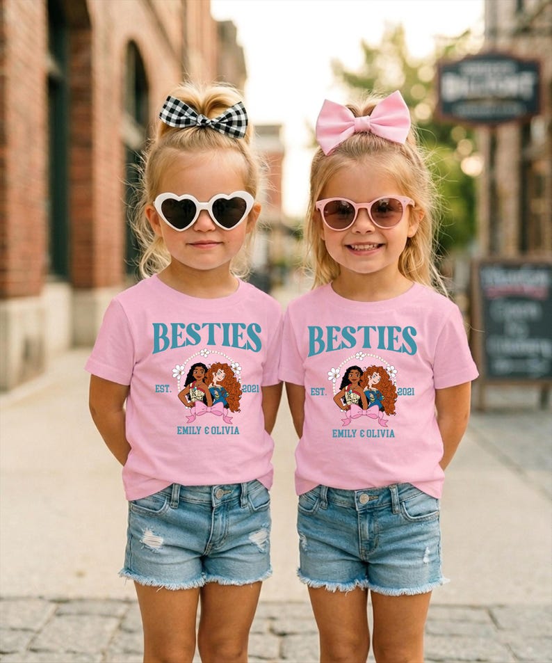 Two young girls wearing matching pink t-shirts with the text "BESTIES" and "EST. 2021 EMILY & OLIVIA" standing side by side on a city street.