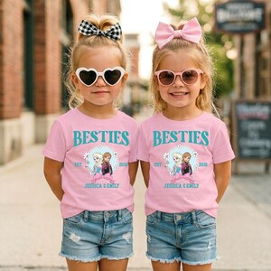 Two young girls wearing matching pink t-shirts with the text "BESTIES" and "EST. 2018 JESSICA & EMILY" printed on them, along with a heart-shaped sun-shaped sunglasses and denim shorts.