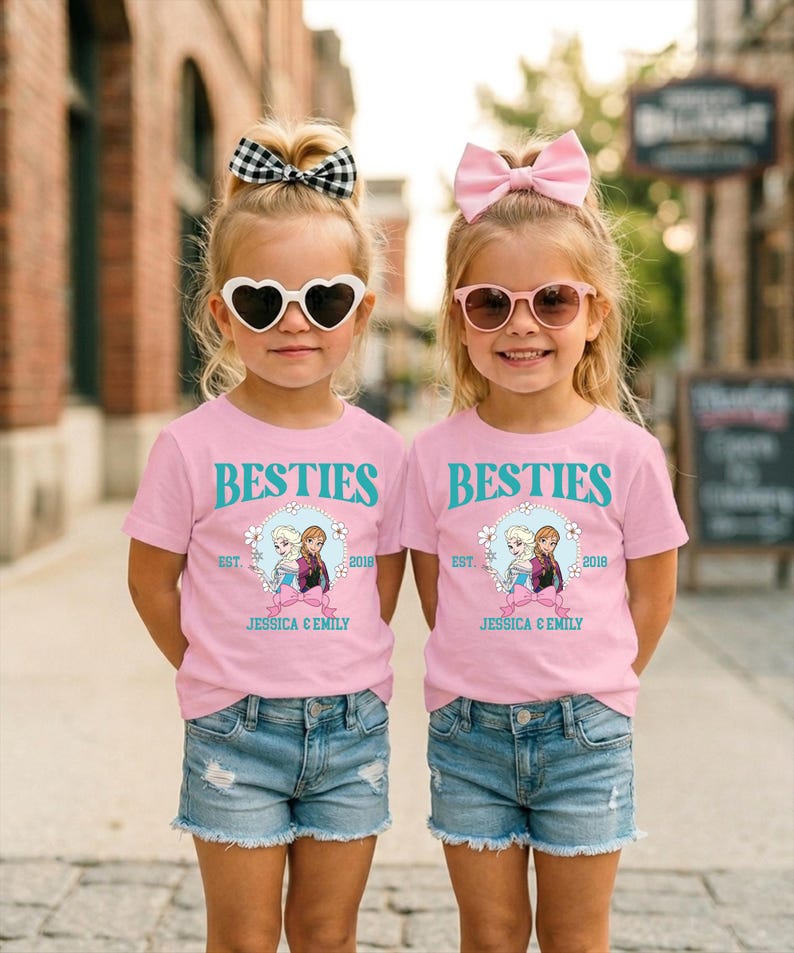 Two young girls wearing matching pink t-shirts with the text "BESTIES" and "EST. 2018 JESSICA & EMILY" printed on them, along with a heart-shaped sun-shaped sunglasses and denim shorts.