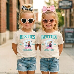 Two young girls wearing matching white t-shirts with the text "BESTIES" and "JESSICA & EMILY" printed on them, along with heart-shaped sunglasses and pink bows, standing together on a city street.