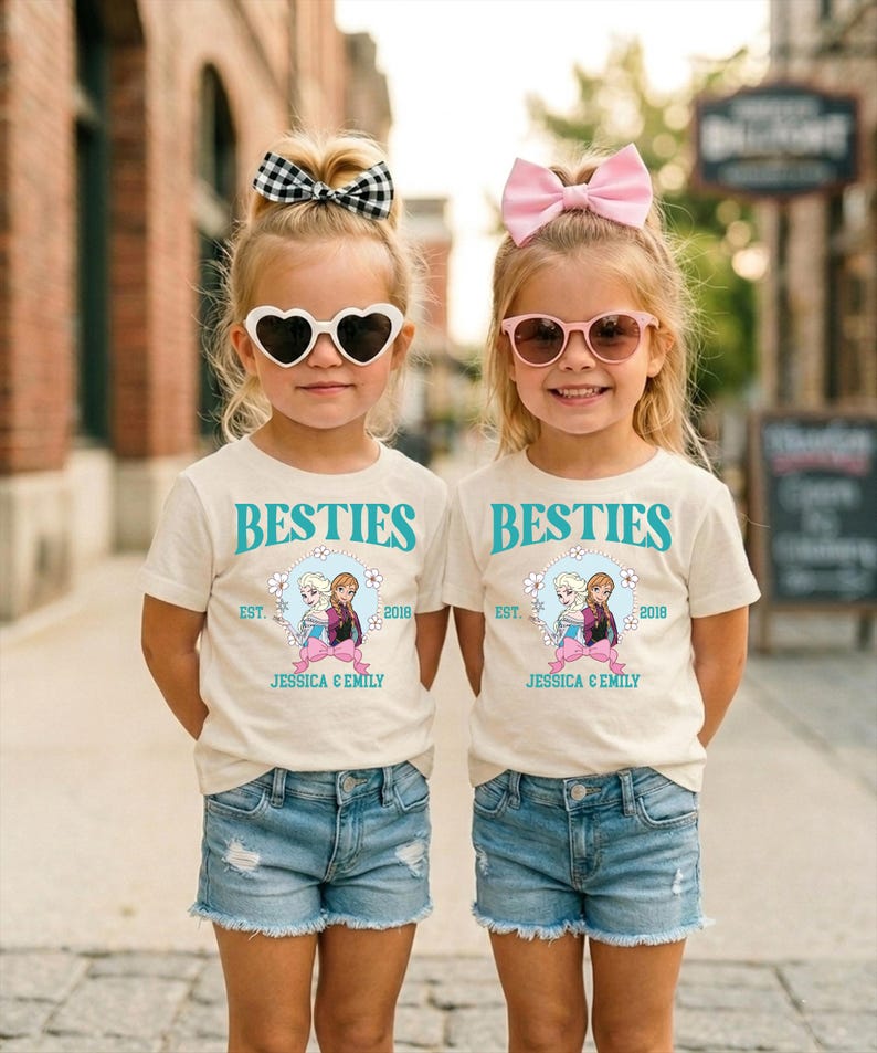 Two young girls wearing matching white t-shirts with the text "BESTIES" and "JESSICA & EMILY" printed on them, along with heart-shaped sunglasses and pink bows, standing together on a city street.