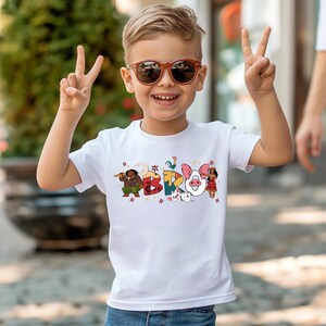 A young boy wearing sunglasses and a white t-shirt with colorful cartoon characters is smiling and making a peace sign with his hands.