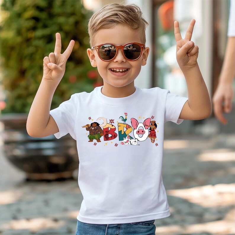 A young boy wearing sunglasses and a white t-shirt with colorful cartoon characters is smiling and making a peace sign with his hands.