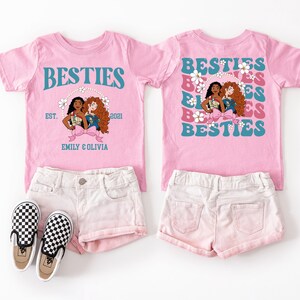 a pink t-shirt with a graphic design featuring three cartoon girls, along with a pair of white shorts and black and white checkered shoes.