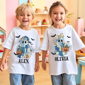 May include: Two children wearing white t-shirts with Halloween ghost designs. The ghost characters are patchwork style, holding jack-o'-lanterns, and surrounded by bats and spiderwebs. The names "ALEX" and "OLIVIA" are printed below the ghosts.