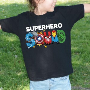 A young child wearing a black t-shirt with the text "SUPERHERO" printed on it, along with various superhero logos and symbols.