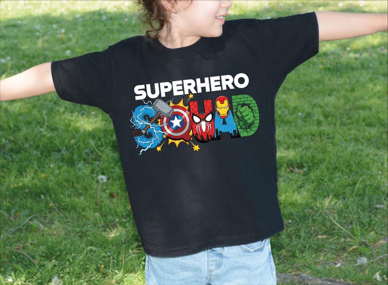 A young child wearing a black t-shirt with the text "SUPERHERO" printed on it, along with various superhero logos and symbols.