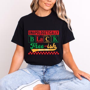 May include: Black t-shirt with the words "UNAPOLOGETICALLY BLACK" in red, green, and yellow, with the words "Free-ish Since 1865" in a stylized font. The shirt has a checkered pattern at the bottom.
