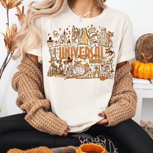 May include: A cream-colored short-sleeve t-shirt featuring a detailed graphic print in shades of orange, brown, and gold. The design displays various Universal Studios landmarks, a globe, and the text "UNIVERSAL STUDIOS" in a stylized font.