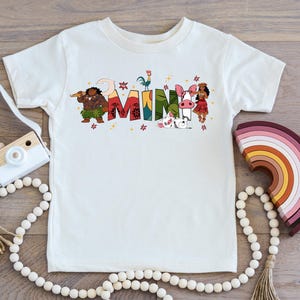 A white t-shirt with colorful text and illustrations, including characters and objects, is displayed on a wooden surface alongside a camera, beads, and a rainbow-colored object.