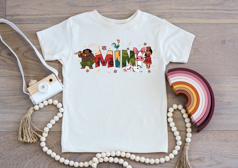 A white t-shirt with colorful text and illustrations, including characters and objects, is displayed on a wooden surface alongside a camera, beads, and a rainbow-colored object.
