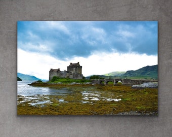 May include: A scenic landscape photograph featuring a stone castle on a small island with a bridge leading to it. The castle is surrounded by green grass and trees, and the water is a calm blue. The sky is cloudy and gray, with hints of blue peeking through.