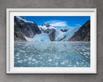 May include: A glacier nestled between snow-capped mountains, with a blue sky and white clouds above. The glacier is reflected in the calm, clear water, which is dotted with small icebergs.