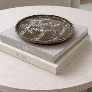 Puede incluir: Un plato decorativo de cerámica marrón con un diseño de puntos blancos descansa sobre dos libros. El libro superior es gris y el inferior es blanco. Los libros están apilados sobre una mesa redonda de color claro.