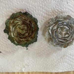 May include: Two decorative, flower-shaped objects. One is a mix of green and brown hues, while the other is a muted gray. Both have a detailed, layered petal design, resembling a rose. They are placed on a white, textured surface.