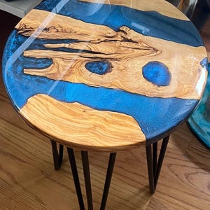Może przedstawiać: Okrągły stolik boczny z drewnianym blatem z niebieskim żywicznym wzorem rzeki. Stół ma naturalny wzór słojów drewna i ciemne metalowe nogi typu hairpin. Średnica około 46 cm.