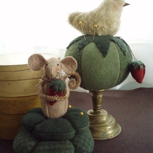 May include: A green pincushion shaped like a tomato with a fluffy white bird perched on top. A smaller green pincushion shaped like a stack of mushrooms with a brown mouse sitting on top. Both pincushions have gold pins.