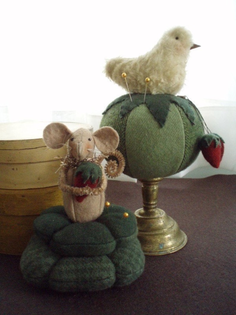 May include: A green pincushion shaped like a tomato with a fluffy white bird perched on top. A smaller green pincushion shaped like a stack of mushrooms with a brown mouse sitting on top. Both pincushions have gold pins.