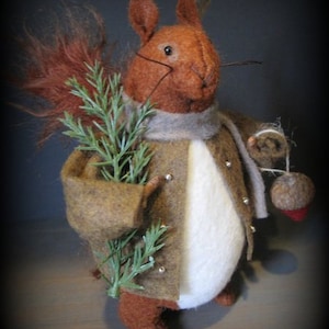 Puede incluir: Una ardilla de fieltro marrón que lleva un abrigo marrón con una bufanda blanca y sostiene una rama de follaje verde. La ardilla sostiene una bellota marrón en su mano derecha.