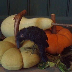 May include: A black crow figurine perched on top of a green and orange pumpkin. The pumpkins are made of felt and have a rustic, handmade look.