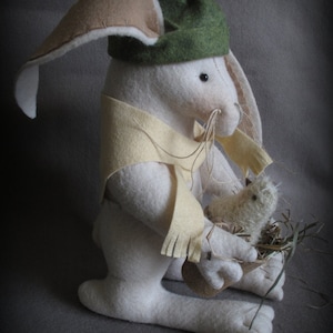 May include: A white felt bunny wearing a green hat with a red pom-pom and a yellow scarf, holding a small white felt bird in a nest.