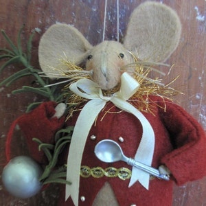 May include: A felt mouse ornament wearing a red coat, a white ribbon, and a gold trim. The mouse is holding a silver spoon and a green sprig.