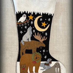 Peut inclure: Une chaussette de Noël en feutre avec un fond noir et des flocons de neige blancs. La chaussette représente une variété d'animaux, dont un loup, un élan, un ours, un phoque et un morse. Les animaux sont empilés les uns sur les autres, le loup en haut et le morse en bas. Il y a une lune croissante et des étoiles en arrière-plan.