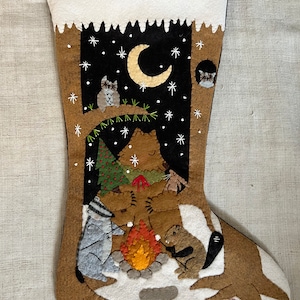 May include: A felt Christmas stocking with a woodland scene. A brown bear, raccoon, and squirrel are gathered around a campfire with a small Christmas tree behind them. Two owls are perched in a tree above them. The stocking is decorated with a crescent moon and stars.