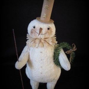May include: A white felt snowman with a brown paper hat decorated with gold stars. The snowman is holding a small green wreath and a brown stick.