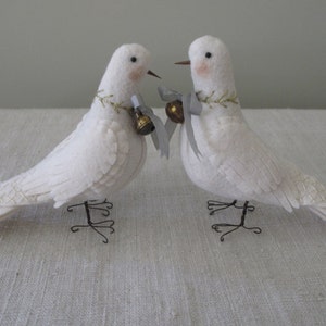 May include: Two white felt doves with gold and silver accents, each with a small bell hanging from their necks. The doves are perched on wire stands.