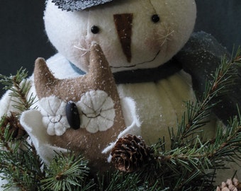 Pode incluir: Um boneco de neve branco usando um chapéu-coco preto e uma echarpe azul segura uma coruja marrom em seus braços. O boneco de neve está rodeado por um galho de pinheiro verde com pinhas marrons.