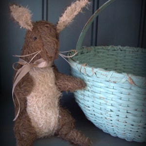 May include: A brown and white plush bunny rabbit with long ears and a beige ribbon around its neck, looking at a light blue wicker basket with a white handle.