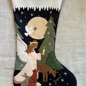 Könnte beinhalten: Schwarzer Weihnachtsstrumpf mit weißem Oberteil und festlichem Design. Das Design umfasst einen Engel, ein Reh, einen Fuchs, ein Kaninchen und eine Taube, mit Vollmond und Schneeflocken. Der Engel hält eine Kerze. Der Strumpf ist aus Filz.