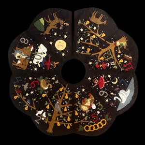 May include: A black Christmas tree skirt with a scalloped edge, featuring embroidered scenes from the Twelve Days of Christmas. The design includes pears, birds, dancers, and other festive elements, with numbers and stars.