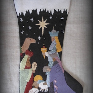 May include: A felt Christmas stocking with a black background and a white snowy top. The stocking features a nativity scene with three wise men, a star, and a camel. The stocking is decorated with white snowflakes.